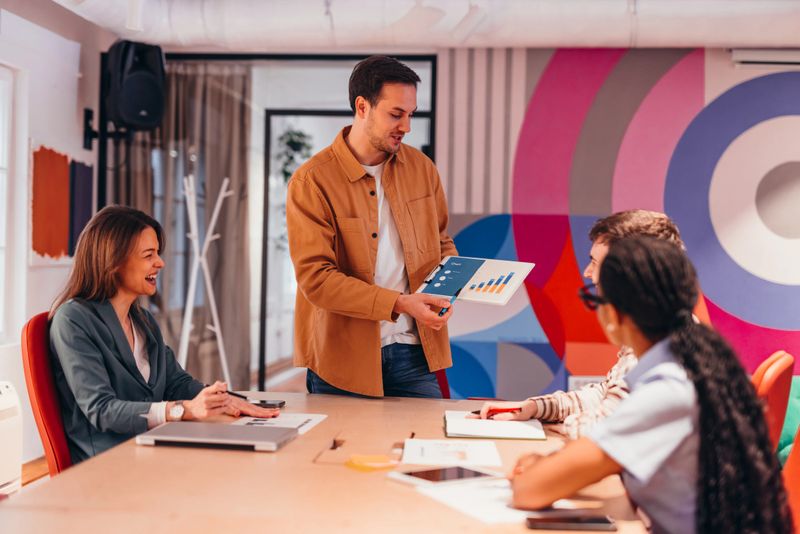 Young manager showing financial charts to his colleagues during a business meeting in a modern office with colorful walls, working together to achieve success