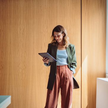 Woman in stylish outfit using a tablet by a wooden wall and window.