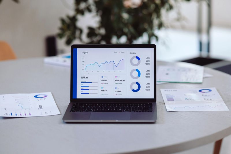 A laptop showcasing data analytics on a desk surrounded by printed reports and greenery. Ideal for illustrating financial, technological, or business-oriented themes in professional settings.