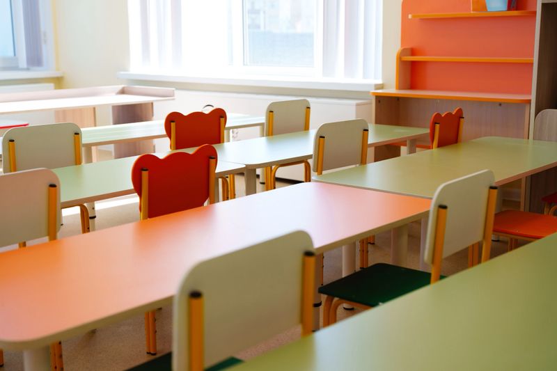 A vibrant classroom features colorful tables and chairs arranged for students. Natural light floods in from large windows, creating an inviting space for learning and collaboration.