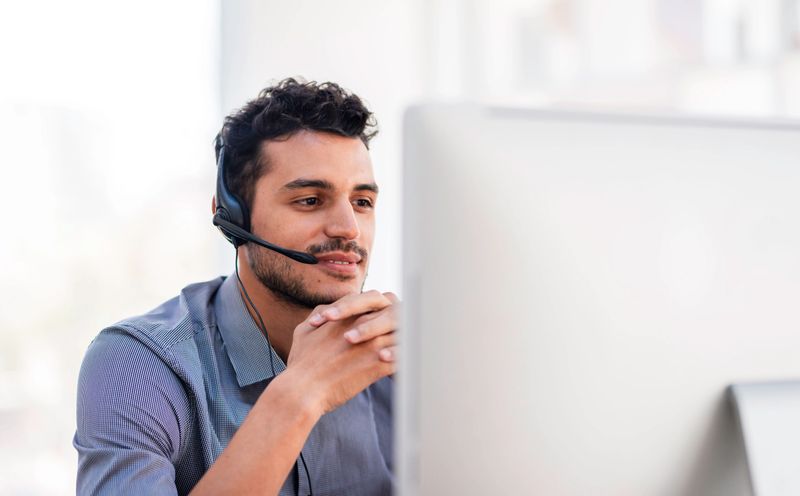 A professional customer service representative in a modern office, wearing a headset and working at a computer. The image portrays professional communication, assistance, and the work environment in customer support.