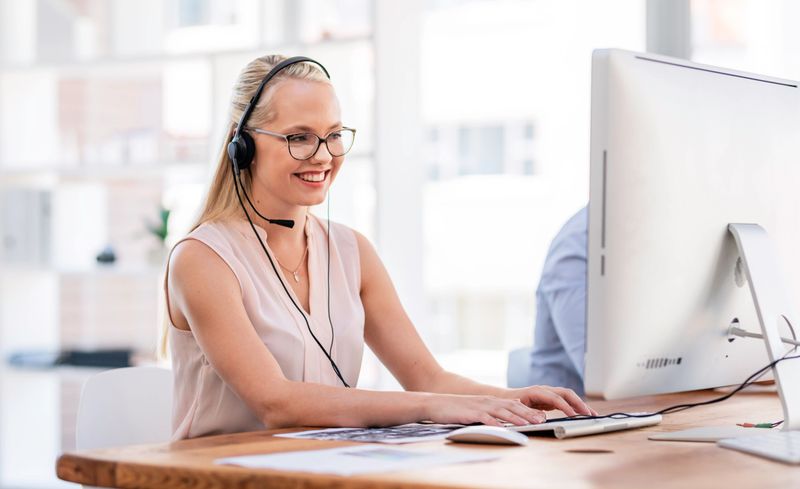 A cheerful customer support employee wearing a headset and glasses, sitting at a desk, typing on a computer. The bright office environment emphasizes professionalism and approachability in customer service settings.
