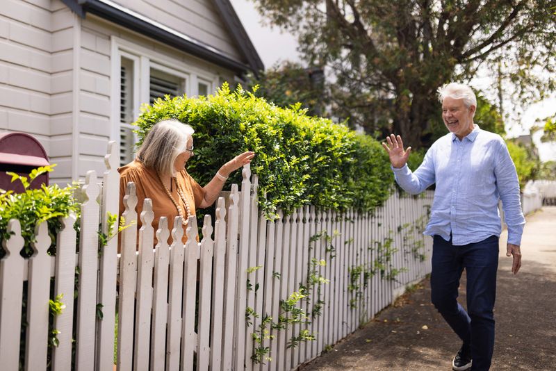 Two cheerful elderly neighbors exchange friendly greetings over a picket fence in a suburban neighborhood, fostering community connections and spreading positivity in a bright and sunny day.