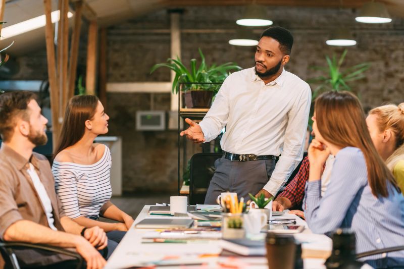 A professional leads a lively discussion with a diverse group in a contemporary workspace. Bright lighting and plants create an inspiring atmosphere for generating ideas and collaboration.