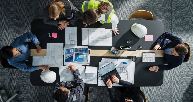 Top Down View of Diverse Team of Real Estate Construction Engineers Working With Blueprints and Documents in an Office. Workspace Of Housing Development Company with Colleagues Using Laptop and Tablet