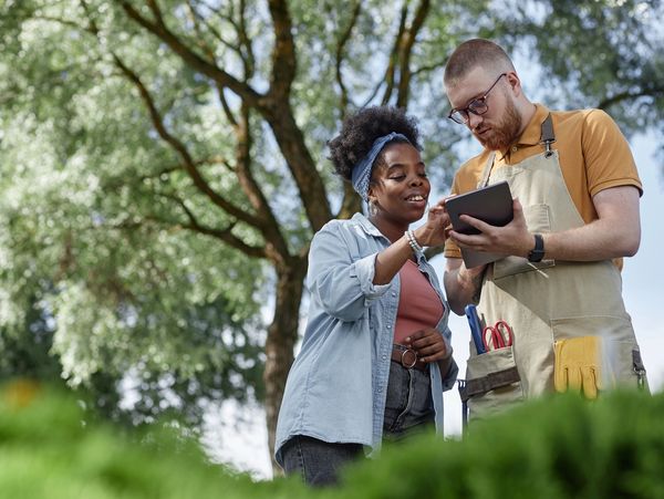 Two gardeners consulting a tablet outdoors under a tree.