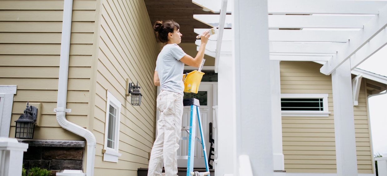 Woman painting white wooden pergola outside a beige house.