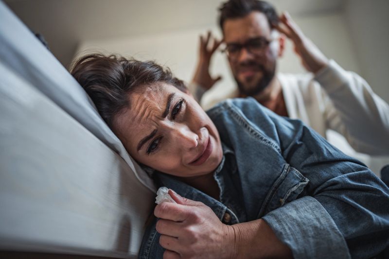 A woman sits on the floor, leaning against the bed, overcome with emotions, tears streaming down her face. Behind her, a man stands, visibly upset.