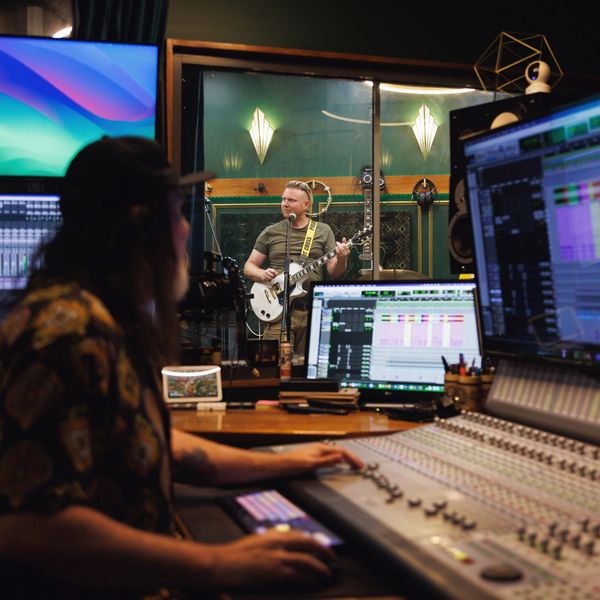 Music producer mixing a track while musician plays guitar in recording studio.