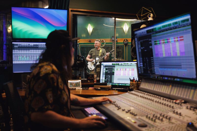 Sound engineer / music producer working in recording studio as the band plays in the rehearsal space