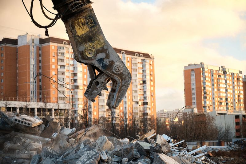 Crusher destroys reinforced concrete materials working at demolition site against apartment buildings at sunset close view