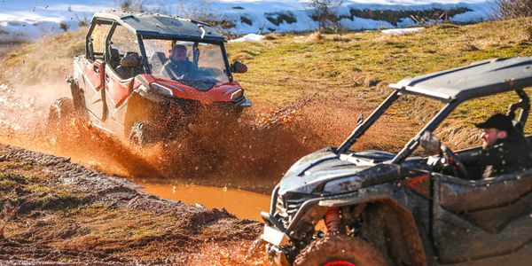 Two off-road vehicles splashing through muddy terrain on a sunny day.