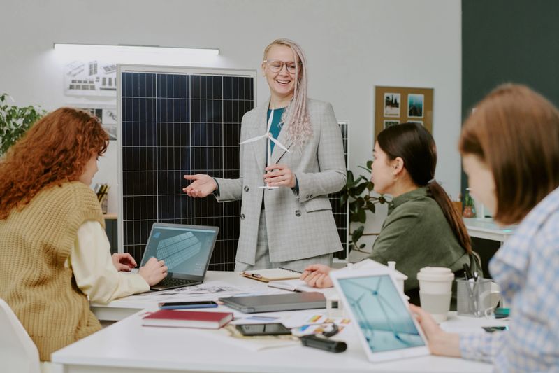 Smiling businesswoman giving presentation in modern office setting with colleagues engaged and discussing at table. Presentation includes solar panel display and various tech devices