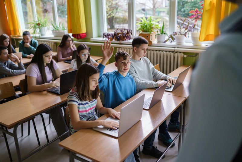 A boy with his hand raised asks a question to the teacher in the classroom. The other children look at the laptop
