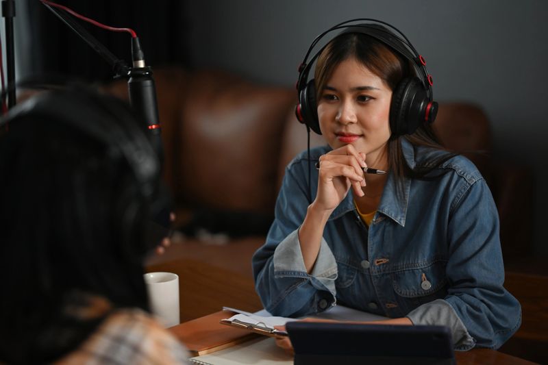 A woman thoughtfully engaging with her guest during a podcast recording, both wearing headphones and using professional microphones in a cozy studio setup.