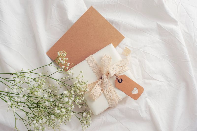 Gift box wrapped with lace ribbon and kraft paper tag with heart cutout next to babys breath flowers on a soft white fabric background. Valentines day concept. Top view, flat lay, mockup.