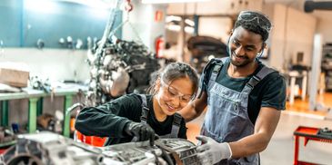 Two mechanics smiling while working on an engine in a garage.