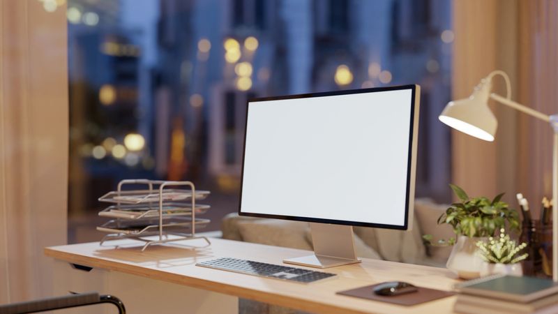 A close-up of a computer with a white screen mockup and decor placed on a wooden table in a modern living room at nighttime, illuminated by warm ambient lighting. 3d render, 3d illustration