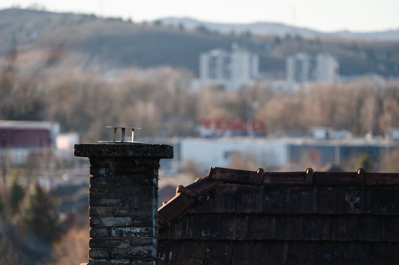 A close-up of a brick chimney and tiled rooftop with an urban skyline and hills in the blurred background. The composition blends rustic architecture with modern city elements.