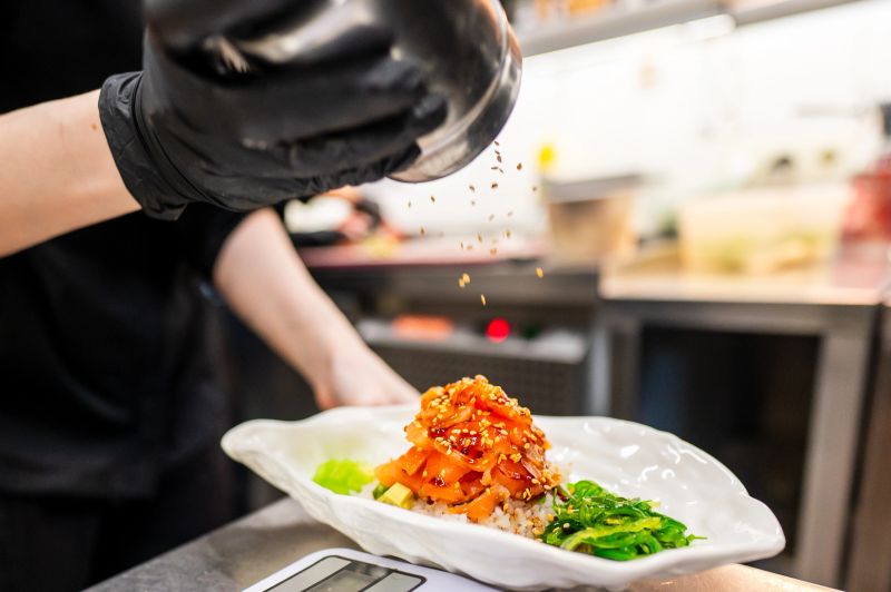 A chef in black gloves sprinkles sesame seeds over a beautifully plated dish featuring vibrant vegetables and fish, showcasing culinary skill in a modern kitchen setting.