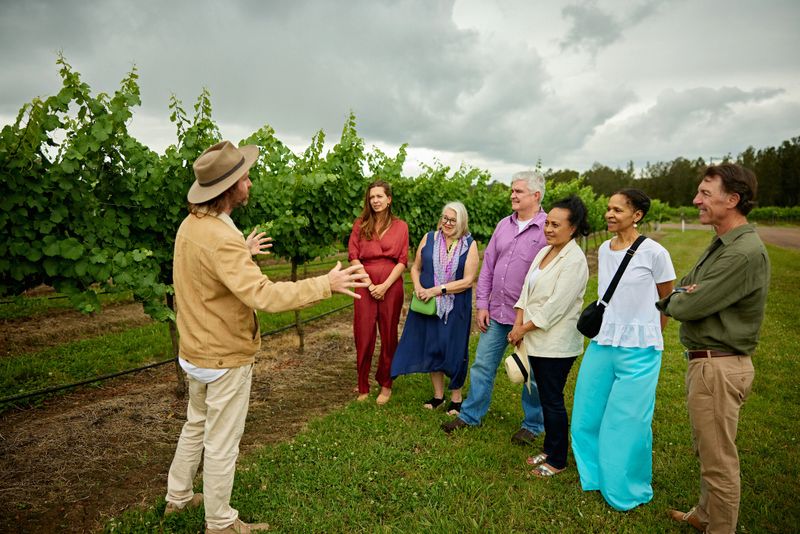 Mid adult man in hat standing outdoors with group of men and women, describing Hunter Valley, Australia’s oldest wine growing region.