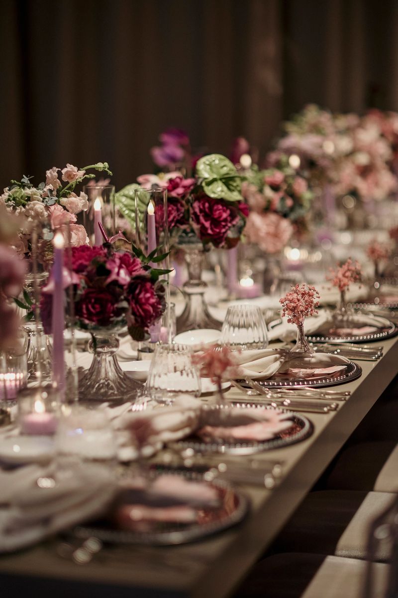 A beautifully decorated wedding reception table adorned with elegant floral arrangements in shades of pink and soft candlelight. The luxurious table setting features delicate glassware, folded napkins, and silver-rimmed plates, creating a warm and romantic atmosphere. Perfect for themes related to weddings, special events, fine dining, and luxury celebrations.