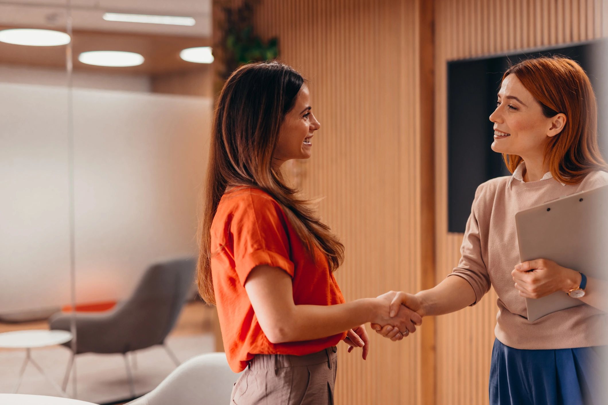 Two women shaking hands and smiling in a modern office setting.