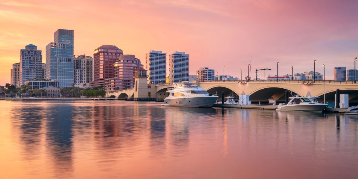 Sunset over a city skyline with boats and a bridge.