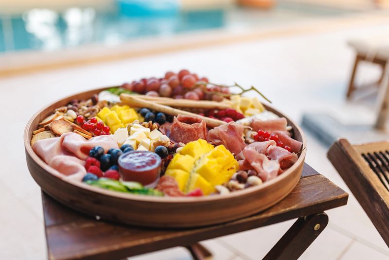 A vibrant charcuterie board filled with assorted fruits, cheeses, and meats on a wooden table by the pool. The setting exudes relaxation, vacation vibes, and culinary enjoyment, perfect for a social gathering or leisure.
