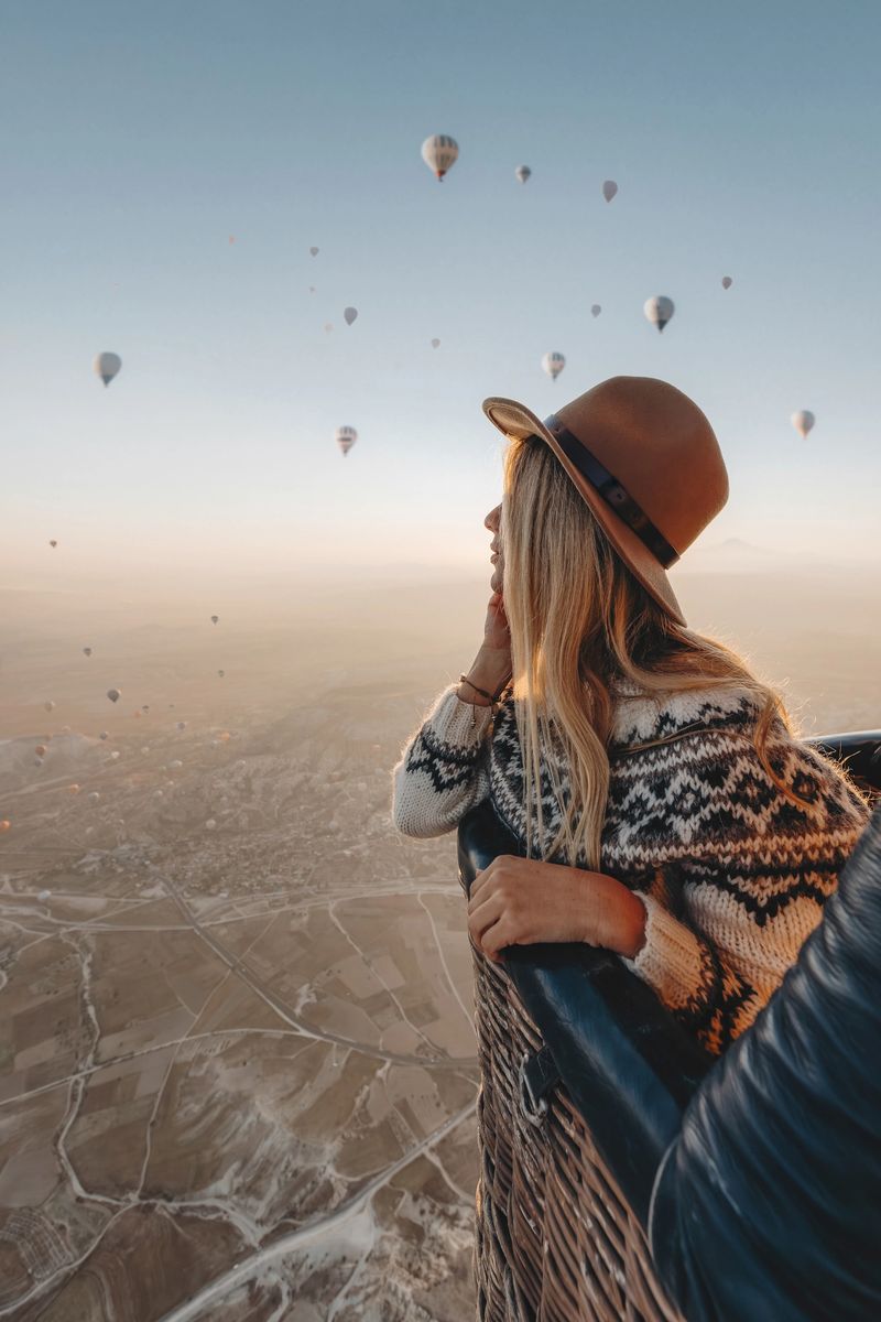 Portrait of s young solo traveler woman with long blonde hair wearing a brown hat and a cozy sweater enjoys a breathtaking hot air balloon ride over Cappadocia, Turkey, at sunrise. The sky is filled with colorful balloons, and the vast landscape below reveals the region’s famous valleys and unique rock formations. This iconic travel experience offers a sense of adventure, freedom, and serenity
