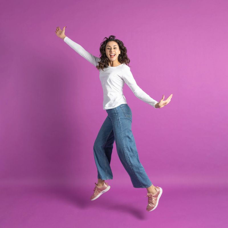 Lifestyle concept. Studio portrait of positive young girl jumping in air against purple background with copy space. Model wearing white blouse, blue jeans and sport shoes