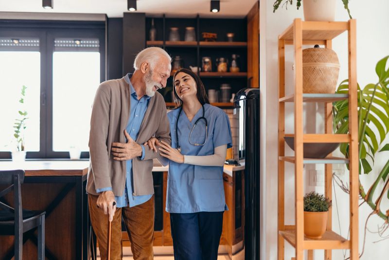 Young nurse assisting a cheerful elderly man walking with a cane in a modern kitchen, delivering compassionate home healthcare and support in a warm, inviting environment