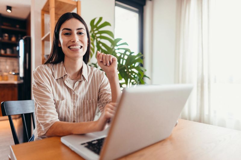 Young woman working from home, sitting at a wooden table in a bright home office, using her laptop, smiling warmly while looking directly at the camera, exuding confidence and positivity