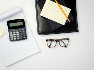 Calculator, glasses, notepad, pencil, and folder on a white desk.