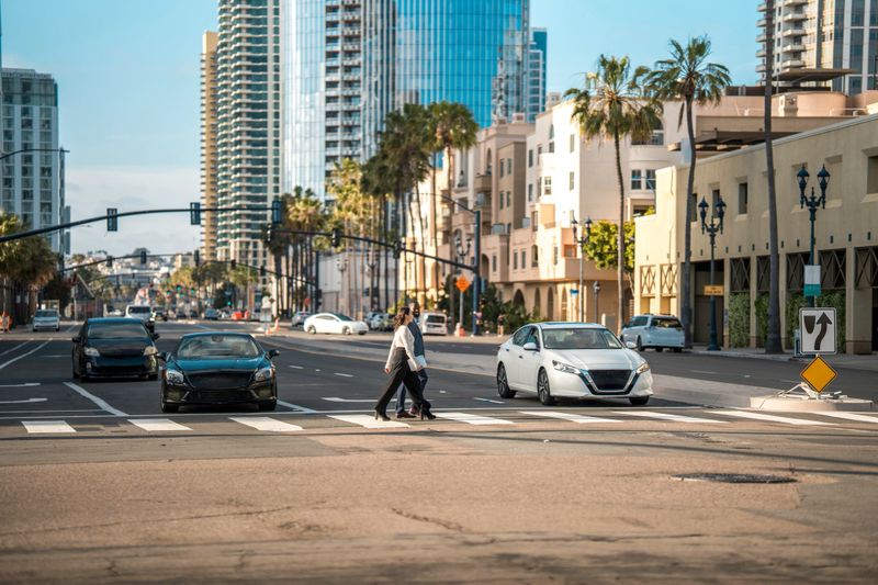 A mid-adult black male and an Asian female executive, both in business attire, walk across a bustling San Diego intersection, with modern skyscrapers lining the backdrop.