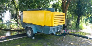 A yellow and gray portable generator trailer parked outdoors under trees.