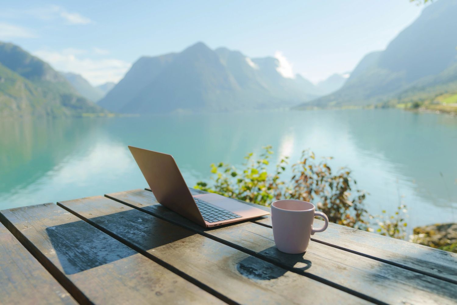 Laptop and coffee on a table overlooking a calm mountain lake.