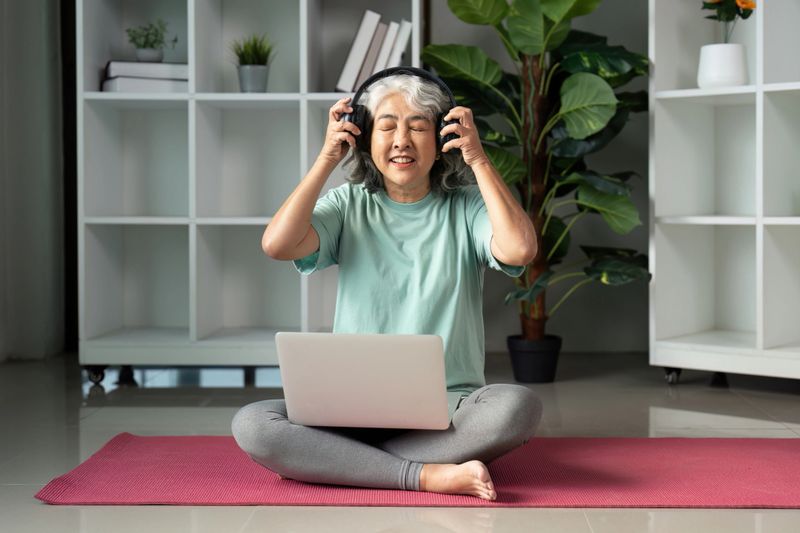 Joyful senior woman sitting cross-legged on a yoga mat, wearing headphones and smiling while engaging with a laptop, promoting a healthy lifestyle.