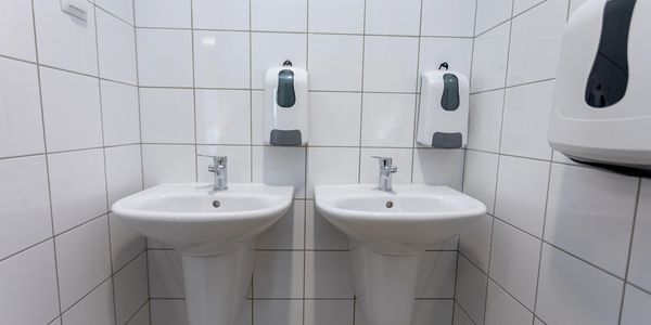 Two white sinks with soap dispensers in a tiled bathroom corner.