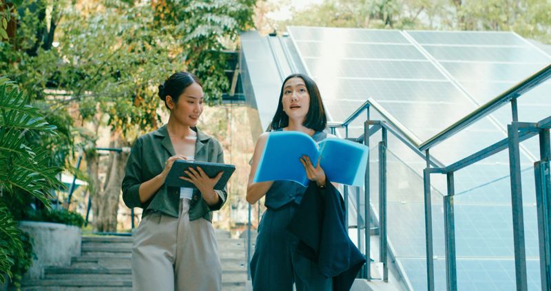 Two young Asian businesswomen digital tablet discuss renewable energy solutions beside staircase equipped with solar panels while walking in urban environment. Sustainable business outdoor concept.