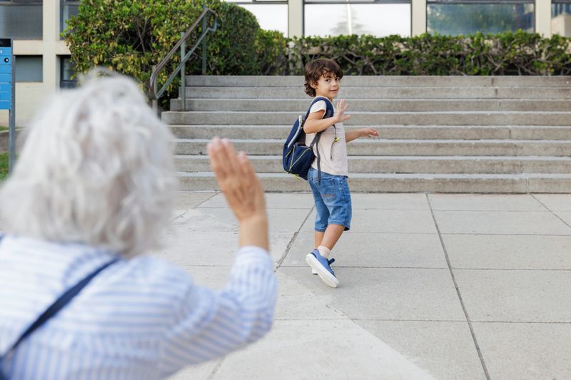 Grandson waving goodbye to his grandmother after school, walking away from her with backpack
