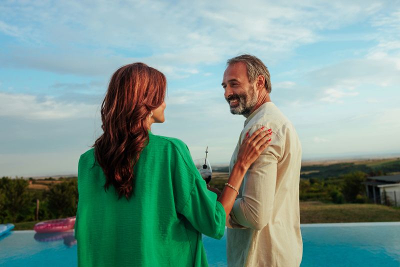 Elegant couple enjoying a sunset by the pool with the woman in a green shirt placing her hand on the shoulder of a middle-aged man in a beige shirt who is smiling.