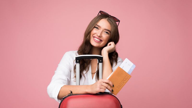 A young woman stands joyfully with a red suitcase and travel documents, conveying excitement for an upcoming journey.