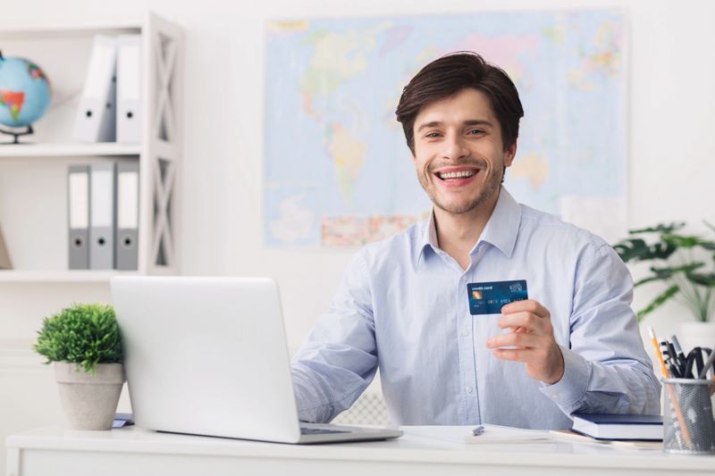 A young man with a cheerful expression holds a credit card in his right hand while sitting at a desk with a laptop. Bright office decor and a world map contribute to a productive atmosphere.