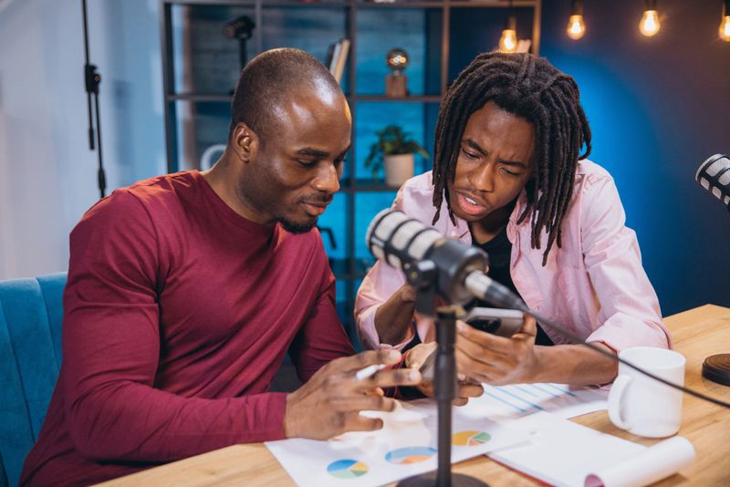 Two African American podcasters analyzing charts on a smartphone while recording their show, engaging in lively discussions and sharing insights