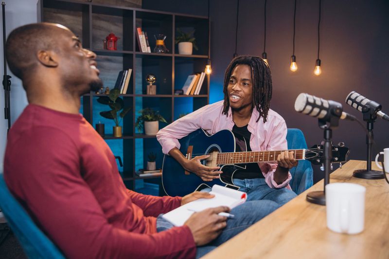 African American acoustic guitarist performing during podcast recording, sitting in professional studio environment with microphone