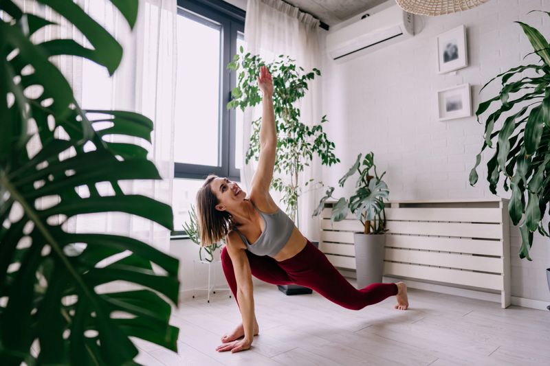 Young woman practicing a revolved crescent lunge yoga pose at home surrounded by houseplants, wearing comfortable sportswear, with a bright window in the background
