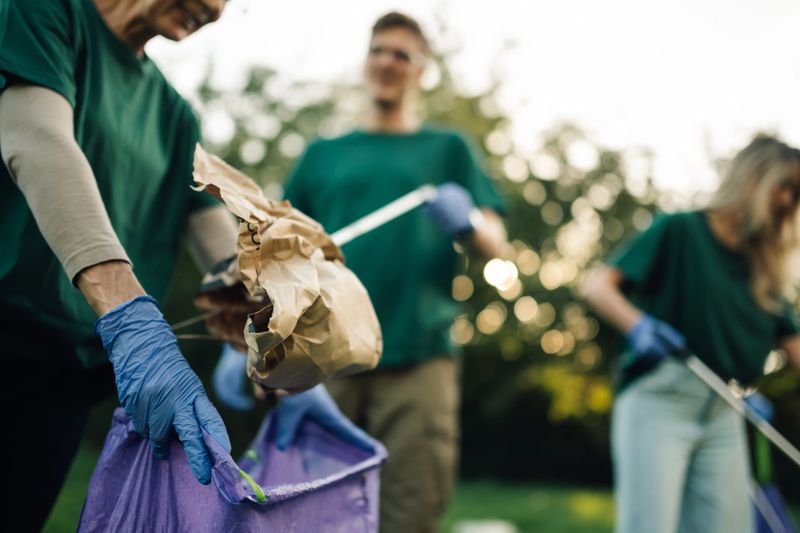 Volunteers wearing green t shirts and gloves are collecting garbage in a public park using trash picker tools and garbage bags