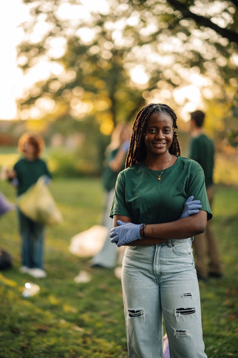 Young woman is posing with her arms crossed, wearing gloves, and smiling while volunteering to clean up a park with a diverse group of people