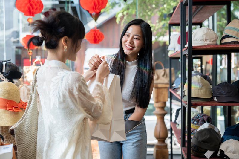 A beautiful, smiling Asian female clothing shop owner or cashier is handing shopping bags to a customer after checkout, creating a pleasant shopping experience in her shop.
